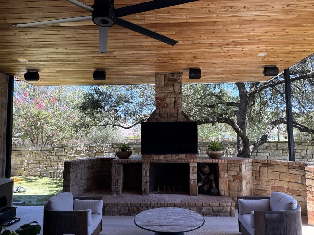 Covered patio with mounted TV, ceiling speakers, and stone fireplace